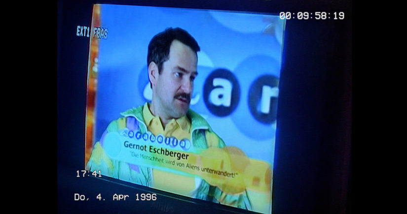 Filmbild aus DER TOTE WINKEL DER WAHRNEHMUNG, das einen TV-Bildschirm zeigt, auf dem ein Mann interviewt wird.